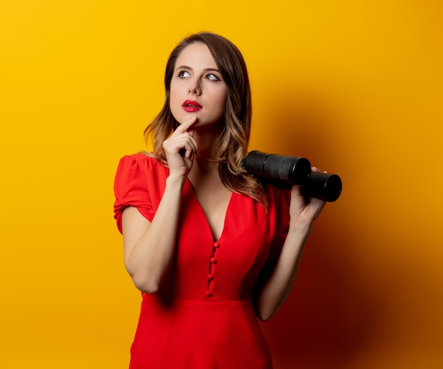 Jeune femme en robe rouge avec des jumelles sur le mur jaune