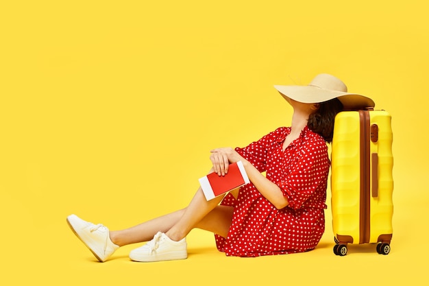 Jeune femme en robe rouge avec valise et passeport en attente de son vol sur fond jaune. retard de vol et voyage annulé