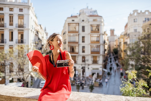 Jeune femme en robe rouge avec ventilateur à main et appareil photo profitant d'une belle vue sur la ville de Valence par temps ensoleillé en Espagne