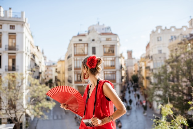 Jeune femme en robe rouge avec ventilateur à main et appareil photo profitant d'une belle vue sur la ville de Valence par temps ensoleillé en Espagne