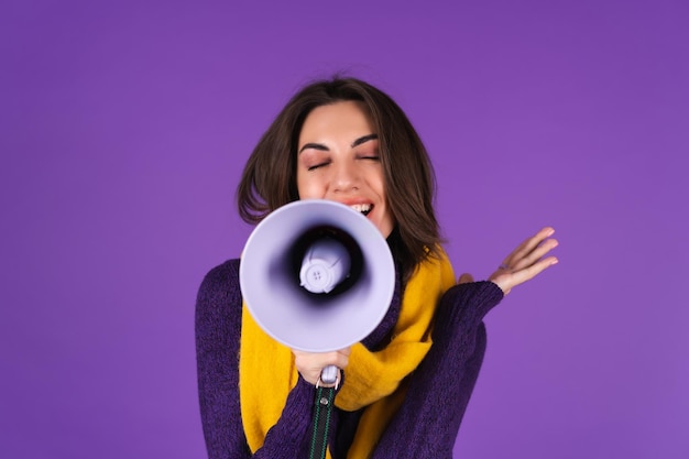 Jeune femme en robe tricotée et écharpe jaune sur fond violet crie joyeusement dans un mégaphone, joyeuse, annonce une vente