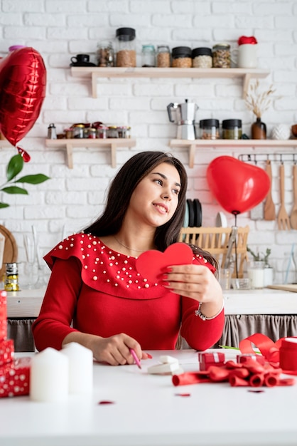 Jeune femme romantique en robe rouge écrit lettre d'amour assis à la cuisine décorée