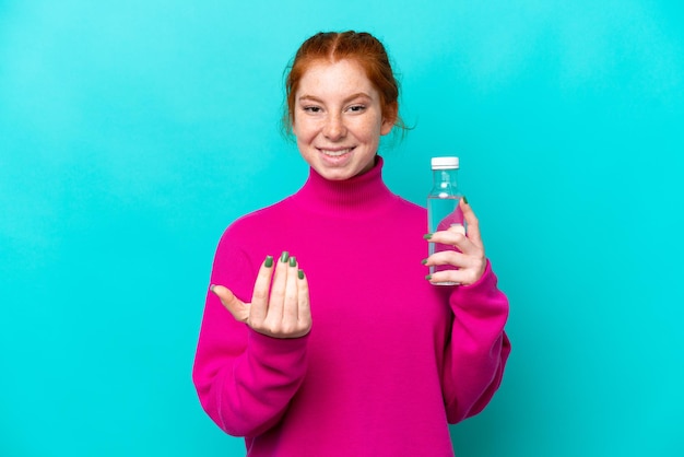 Jeune femme rougeâtre caucasienne avec une bouteille d'eau isolée sur fond bleu invitant à venir avec la main Heureux que vous soyez venu