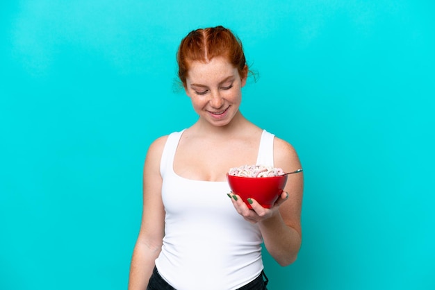 Jeune femme rougeâtre tenant un bol de céréales isolé sur fond bleu avec une expression heureuse