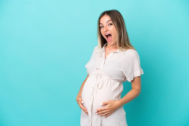 Jeune femme roumaine isolée sur fond bleu enceinte et faisant un geste surprise