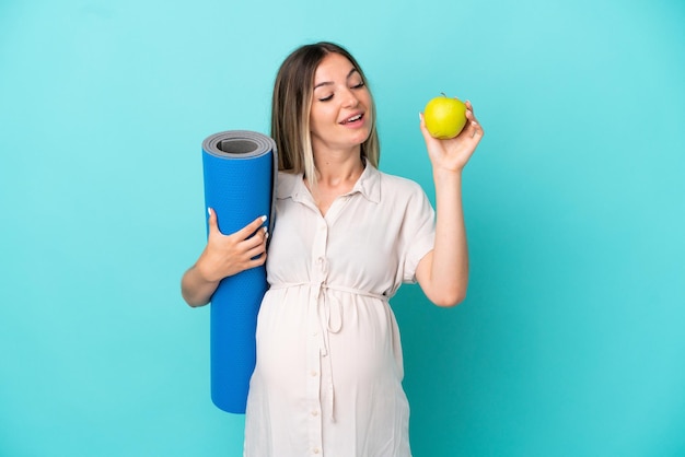 Jeune femme roumaine isolée sur fond bleu enceinte tenant une pomme et allant à des cours de yoga