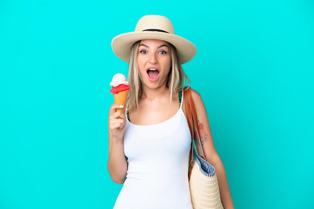 Jeune femme roumaine tenant une crème glacée et un sac de plage isolé sur fond bleu avec une expression faciale surprise et choquée