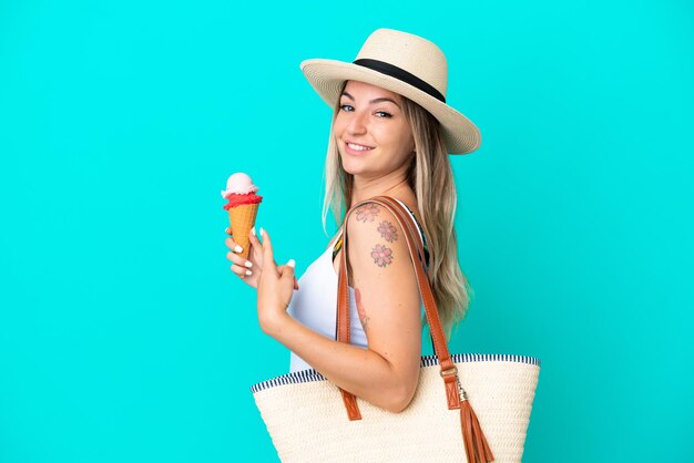 Jeune femme roumaine tenant une crème glacée et un sac de plage isolé sur fond bleu et le pointant