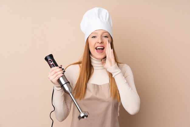 Jeune femme rousse à l'aide d'un mélangeur à main criant avec la bouche grande ouverte