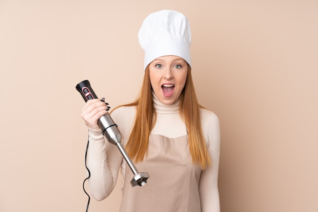 Jeune femme rousse à l'aide d'un mélangeur à main avec surprise et expression faciale choquée