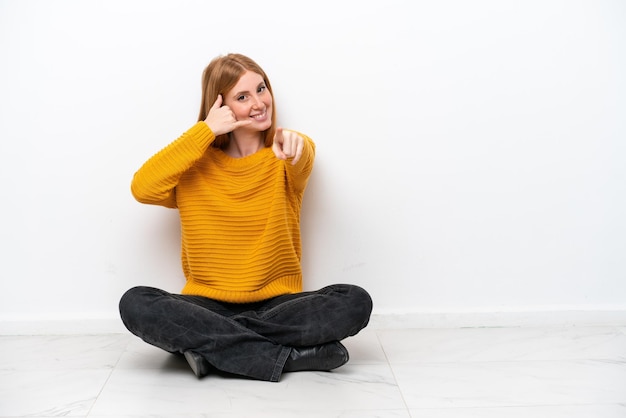 Jeune femme rousse assise sur le sol isolé sur fond blanc faisant un geste de téléphone et pointant vers l'avant