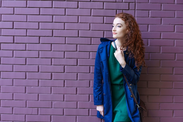 Jeune femme rousse attirante réfléchie dans l'imperméable bleu regardant l'appareil-photo