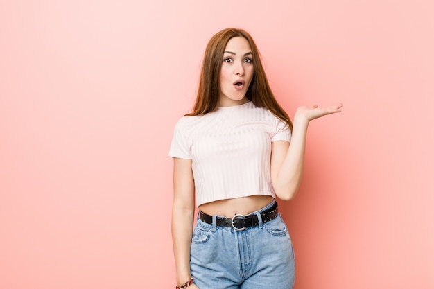 Jeune femme rousse au gingembre contre un mur rose impressionné tenant la paume de la surface.