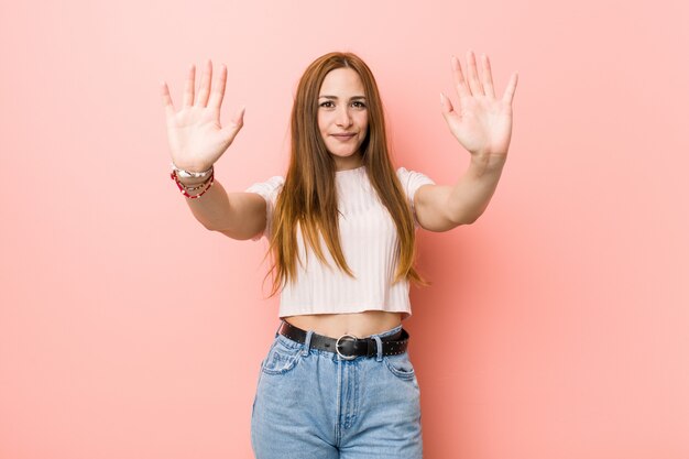 Jeune femme rousse au gingembre contre un mur rose montrant le numéro dix avec les mains.