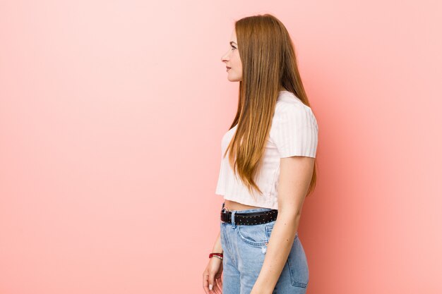 Jeune femme rousse au gingembre contre un mur rose regardant à gauche, pose sur le côté.