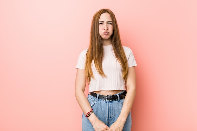 Jeune femme rousse au gingembre contre un mur rose souffle les joues, a l'air fatigué