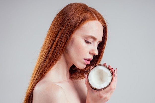 Jeune femme rousse au gingembre avec une peau idéale et une oreille rousse parfaite tenant la noix de coco dans les mains en fond de studio blanc.