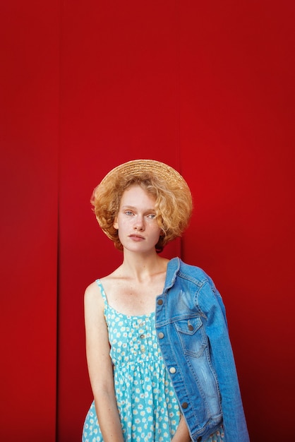 Jeune femme rousse bouclée en chapeau de paille bleu robe d'été et veste jeans debout sur backgrou rouge foncé