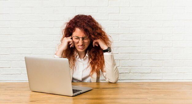 Jeune femme rousse bouclée travaillant avec son ordinateur portable couvrant les oreilles avec les mains.