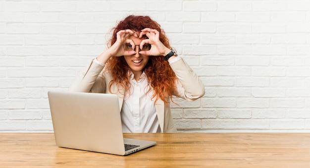 Jeune femme rousse bouclée travaillant avec son ordinateur portable montrant un signe correct sur les yeux