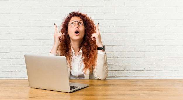 Jeune femme rousse bouclée travaillant avec son ordinateur portable pointant à l'envers avec la bouche ouverte.