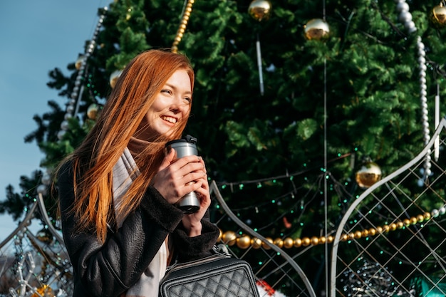 Jeune femme rousse buvant du café errant près de l'arbre de noël dans les rues de la ville d'hiver