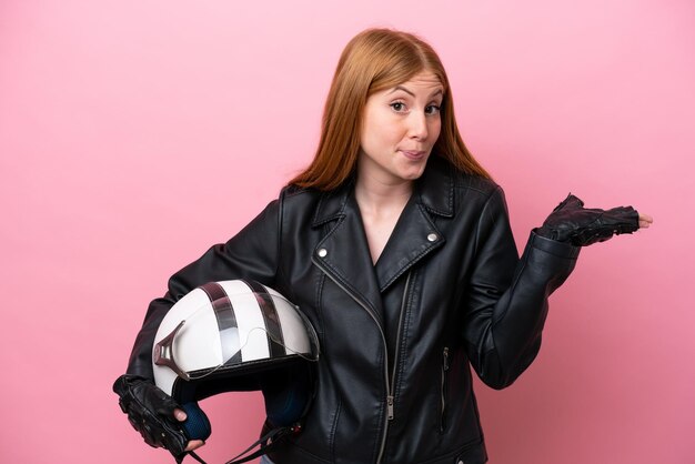 Jeune femme rousse avec un casque de moto isolé sur fond rose ayant des doutes tout en levant les mains