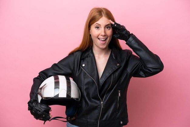 Jeune femme rousse avec un casque de moto isolé sur fond rose avec une expression surprise