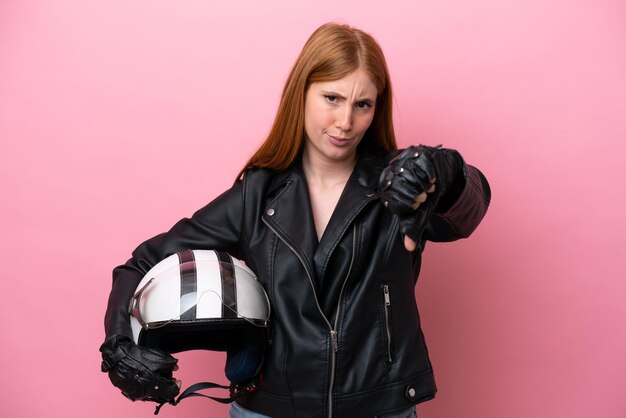 Jeune femme rousse avec un casque de moto isolé sur fond rose montrant le pouce vers le bas avec une expression négative