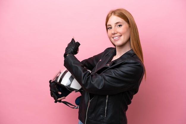 Jeune femme rousse avec un casque de moto isolé sur fond rose pointant vers l'arrière