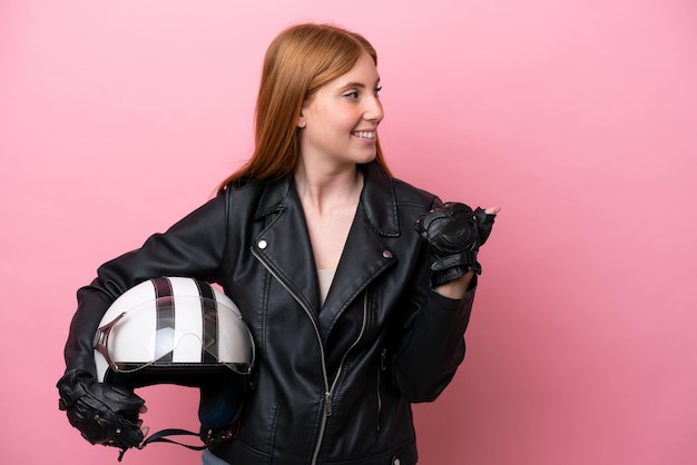 Jeune femme rousse avec un casque de moto isolé sur fond rose pointant vers le côté pour présenter un produit