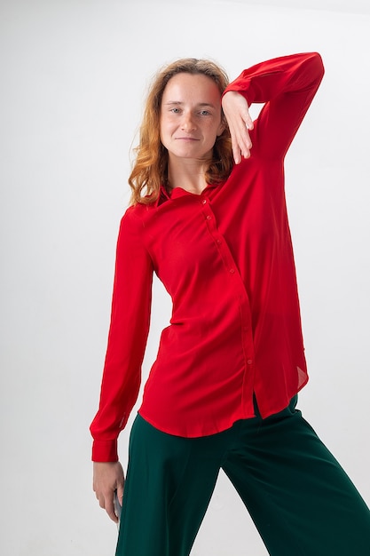 Jeune femme rousse caucasienne aux cheveux longs en chemise rouge isolée sur fond blanc exhibant, tenant la main près de son visage. femme drôle s'amuser dans la pose de danse disco