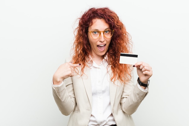 Jeune femme rousse caucasienne tenant une carte de crédit surpris en se montrant, souriant largement.