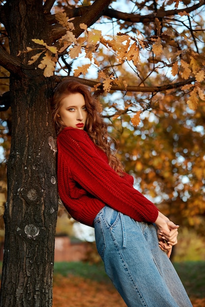 Jeune femme rousse dans un pull rouge se promène dans le parc. Portrait de beauté automne d'une femme rousse à la mode au coucher du soleil