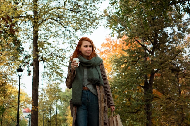 Jeune femme rousse élégante en manteau avec des sacs à provisions artisanaux marchant dans le parc de la ville à l'automne