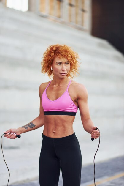 Jeune femme rousse européenne en vêtements sportifs sautant avec une corde à l'extérieur