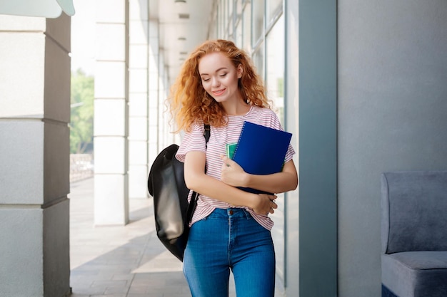 Jeune femme rousse à l'extérieur sur le chemin des cours. Jeune fille souriante tenant des cahiers. Éducation et entrée dans le concept universitaire