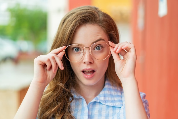 Jeune femme rousse à l'extérieur avec des lunettes et une expression surprise
