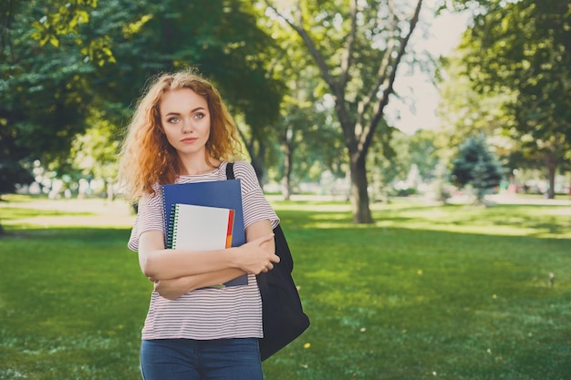 Jeune femme rousse à l'extérieur. Étudiante debout dans le parc avec un cahier d'exercices et un sac à dos. Notion d'éducation