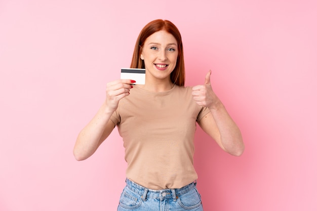 Jeune femme rousse sur fond rose isolé, détenant une carte de crédit