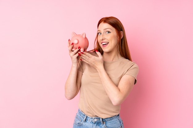 Jeune femme rousse sur fond rose isolé tenant une grande tirelire