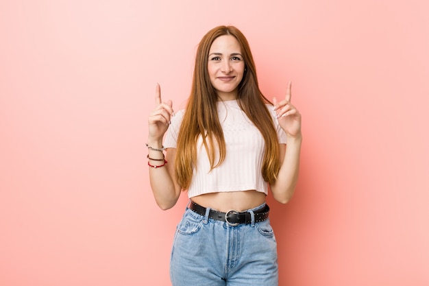 Jeune femme rousse gingembre contre un mur rose indique avec les deux doigts avant montrant un espace vide.