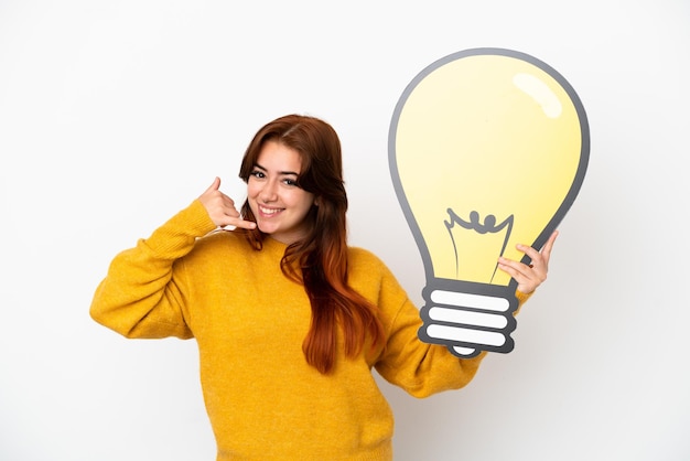 Photo jeune femme rousse isolée sur fond blanc tenant une icône d'ampoule et faisant un geste de téléphone