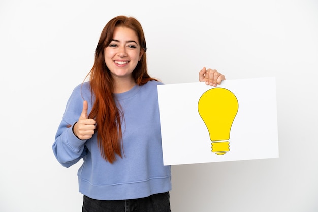 Jeune femme rousse isolée sur fond blanc tenant une pancarte avec l'icône de l'ampoule avec le pouce vers le haut