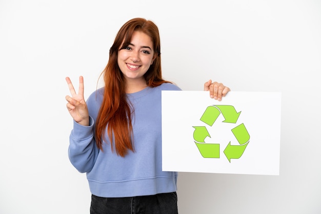 Jeune femme rousse isolée sur fond blanc tenant une pancarte avec icône de recyclage et célébrant une victoire