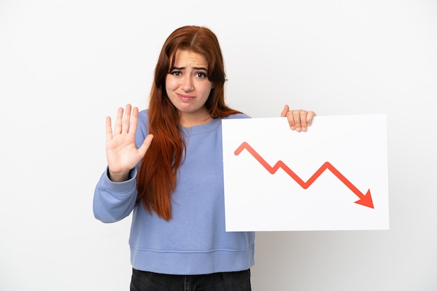 Jeune femme rousse isolée sur fond blanc tenant une pancarte avec un symbole de flèche de statistiques décroissantes et faisant un panneau d'arrêt