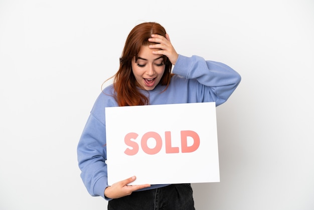 Jeune femme rousse isolée sur fond blanc tenant une pancarte avec texte VENDU avec expression surprise