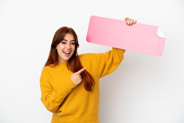 Jeune femme rousse isolée sur fond blanc tenant une pancarte vide et la pointant