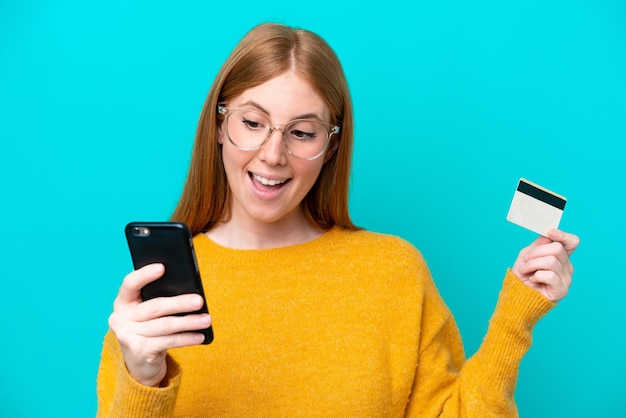 Jeune femme rousse isolée sur fond bleu achetant avec le mobile avec une carte de crédit