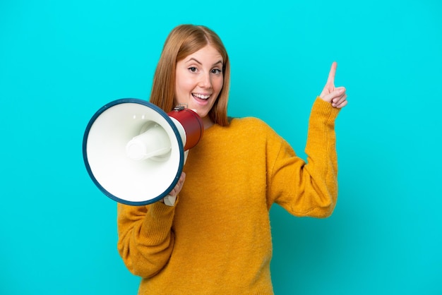 Jeune femme rousse isolée sur fond bleu criant à travers un mégaphone et pointant du côté